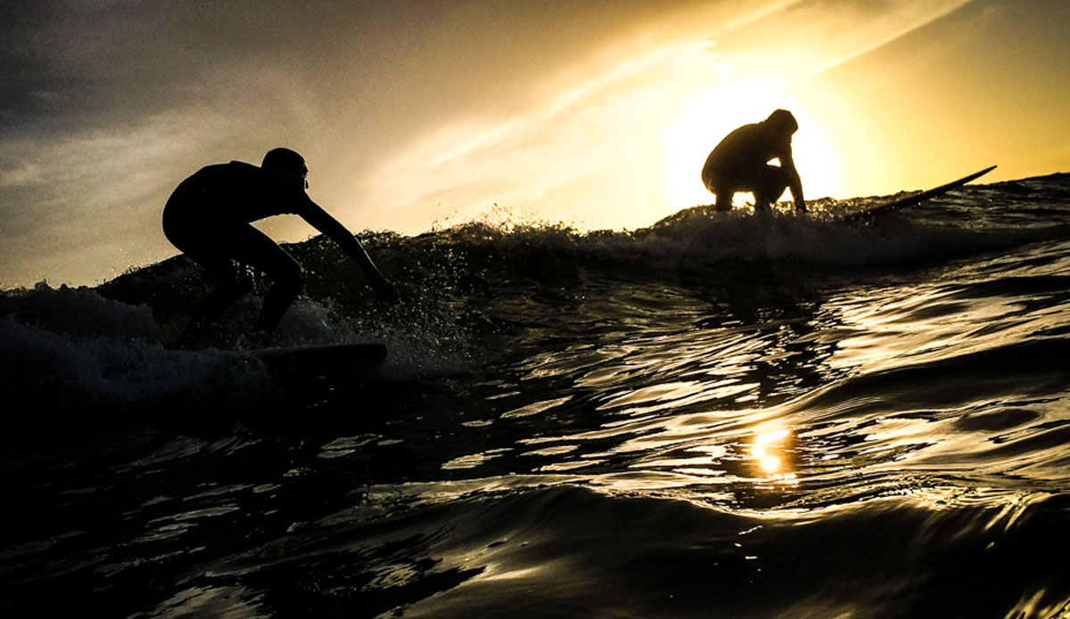 Two surfers drop in on a wave together, perfectly silhouetted against the setting sun. This time of day is called the golden hour for a reason. Photo: <a href=\"https://richbrooksphotography.squarespace.com/\"> Rich Brooks</a>