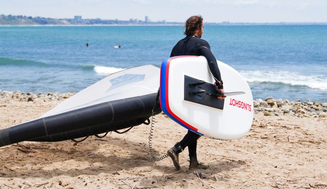 a man carrying the slingshot ltf board from the beach to the water with a wing