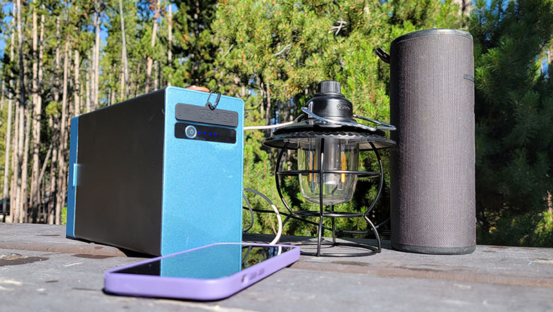 the FJD Pony 500 portable power station charging an iphone, lantern, and bluetooth speaker at a campground in the forest