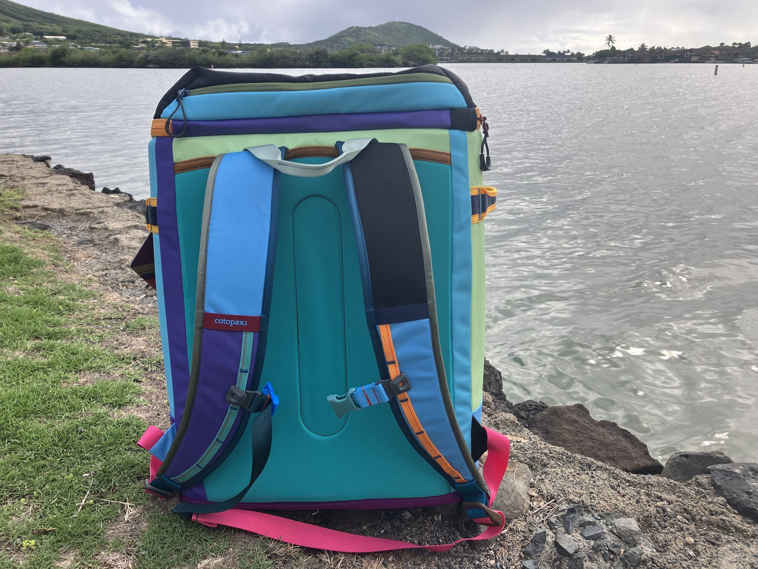 a Cotopaxi backpack cooler sitting on the grass near the water