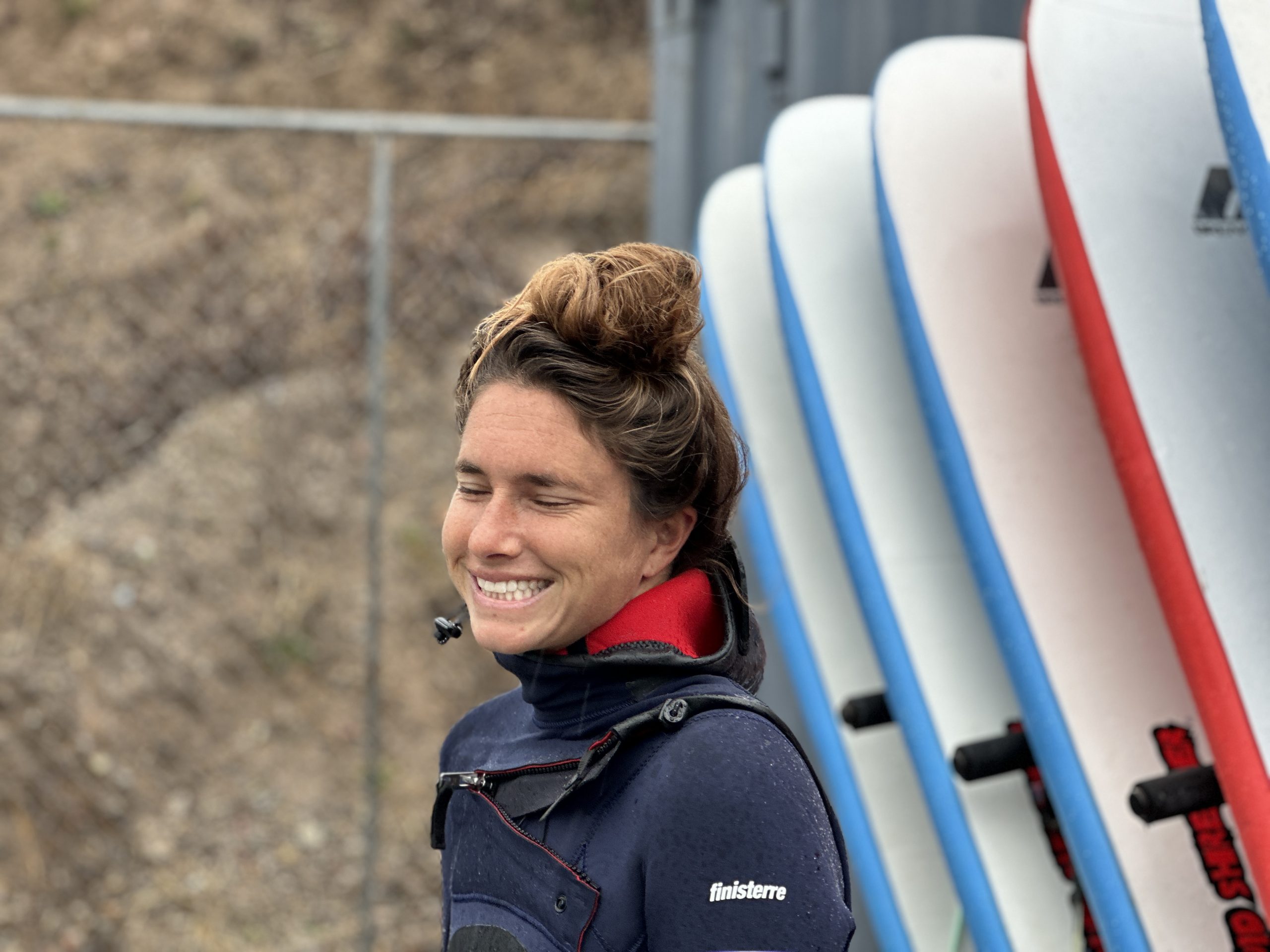 a woman smiling in front of surfbaords in a wetsuit