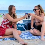 two women with soft coolers drinking a beverage on the beach