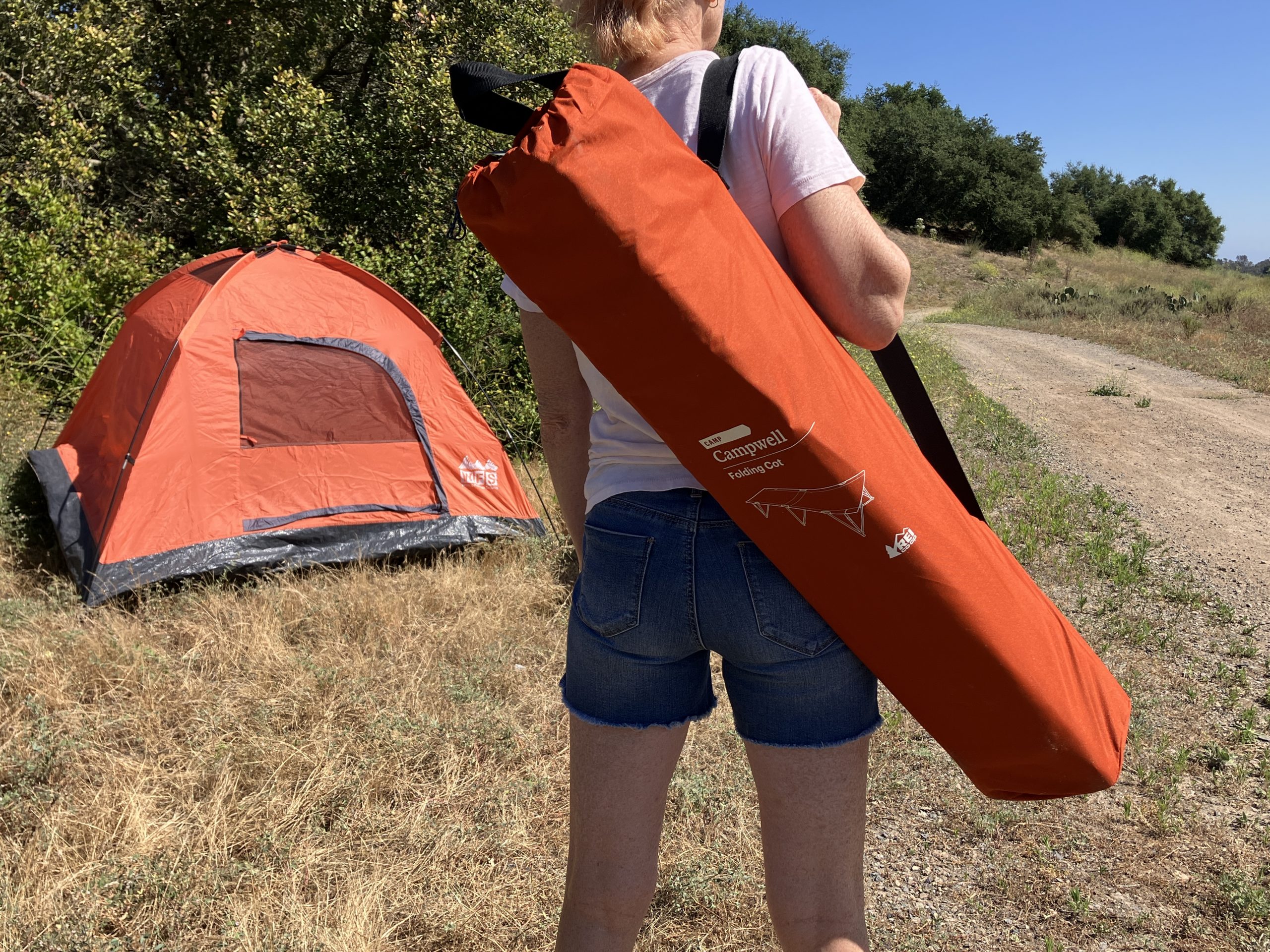 a woman carrying a camping cot folded up in front of a tent