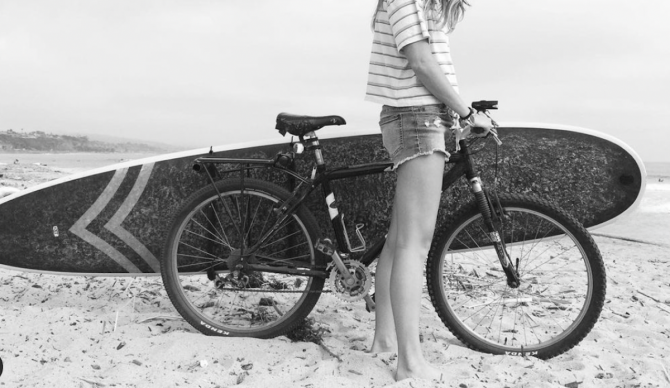 Woman standing with a surfboard on a bike rack at the beach